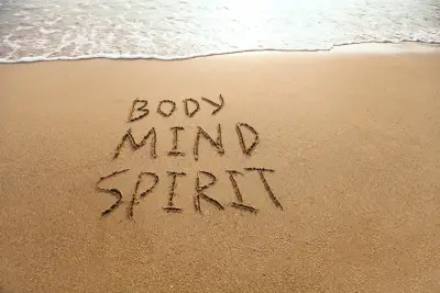 Body-Mind-Spirit handwritten words on the sand, showing body mind spirit in bold letters, with waves coming in